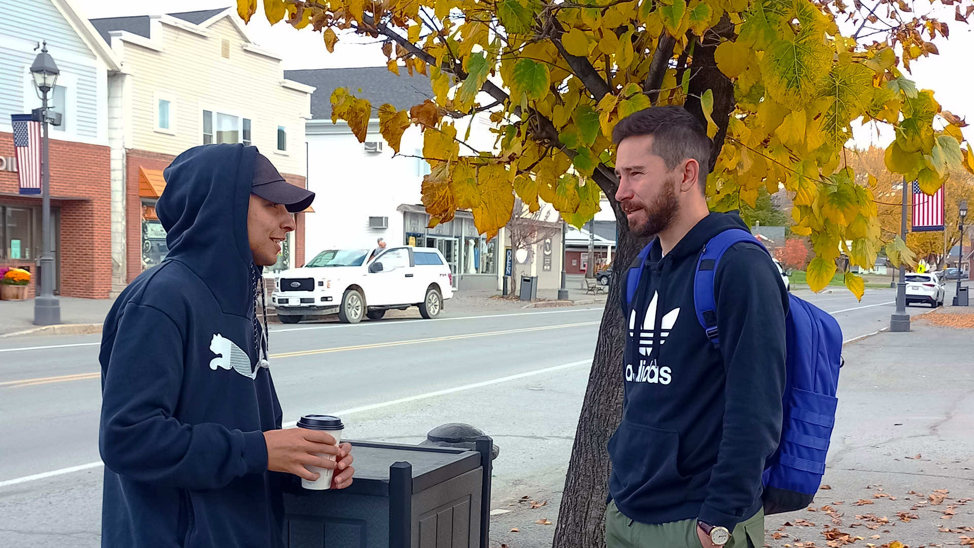 Two students have a conversation while standing on the sidewalk in downtown Fort Kent