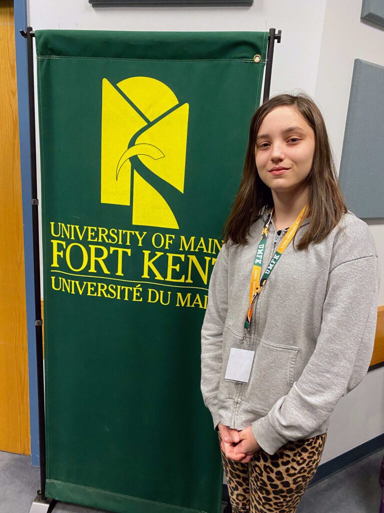 County spelling bee champion Claire Thorsnes standing next to a University of Maine at Fort Kent banner